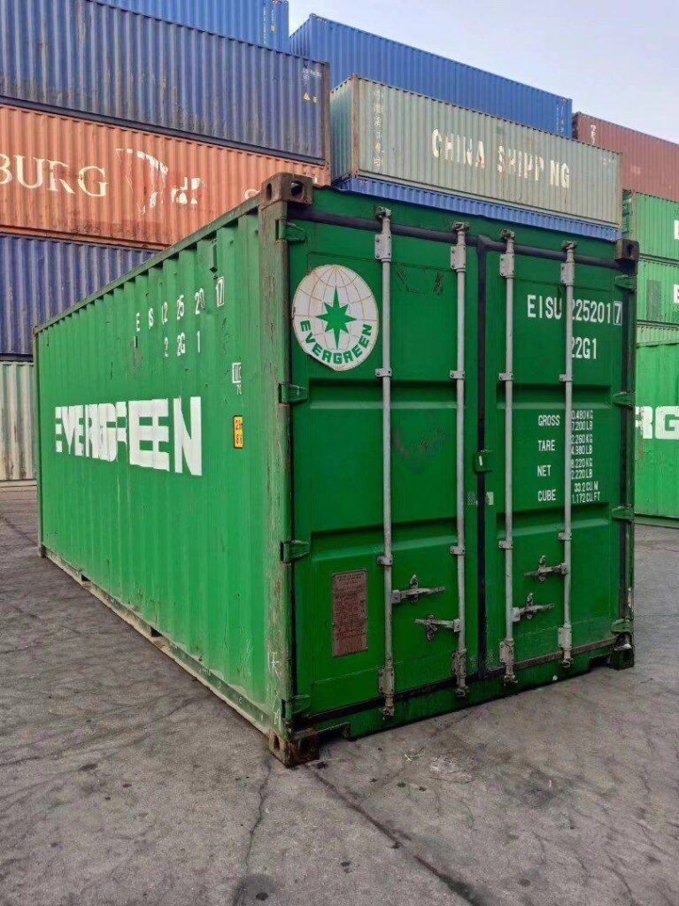 Grüner 20-Fuß-Container in Schrägansicht vor gestapelten Seecontainern im Depot