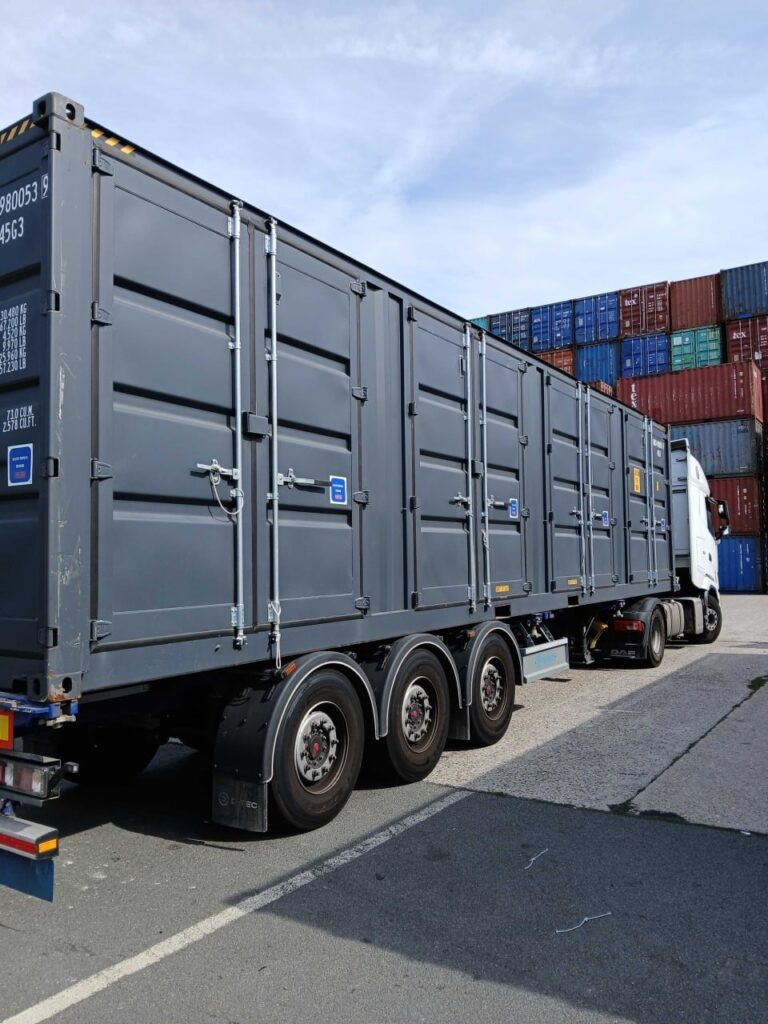Dunkelgrauer 40-Fuß-High-Cube-Container mit Seitentüren auf einem Sattelauflieger vor gestapelten Containern