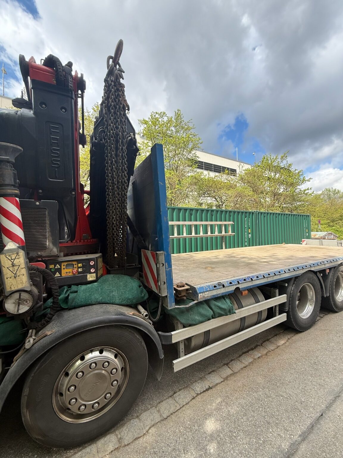 Lastwagen mit Ladekran und leerer Pritsche, dahinter ein grüner Container